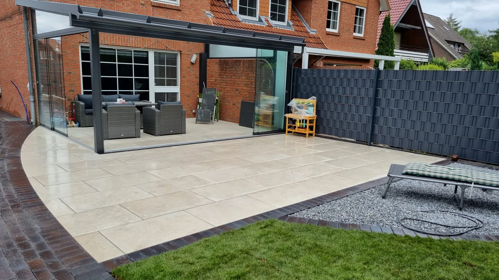 Überdachte Terrasse aus hellen Keramikplatten mit Sitzgruppe und Rasenkante